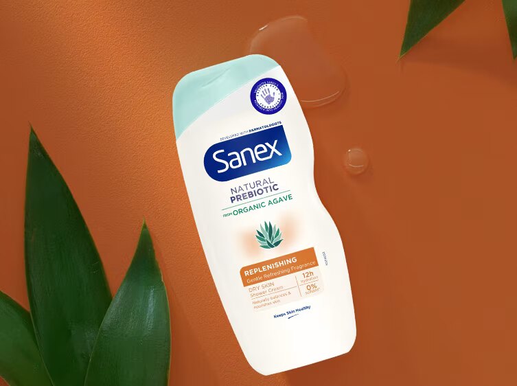 Flacon de la crème de douche Sanex Natural Prebiotic Replenishing, posé à plat sur un fond orange avec des feuilles vertes et des gouttes de gel.