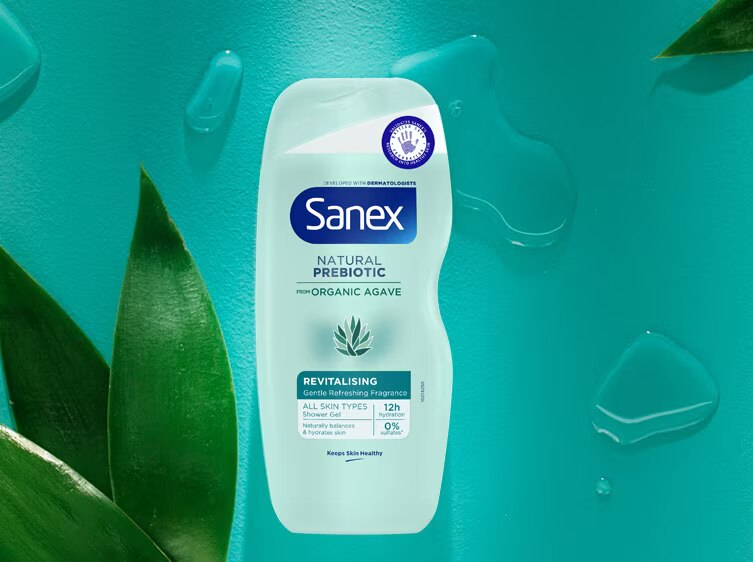 Flacon du gel douche Sanex Natural Prebiotic Revitalising, posé à plat sur un fond turquoise avec des feuilles vertes et des gouttes d'eau.