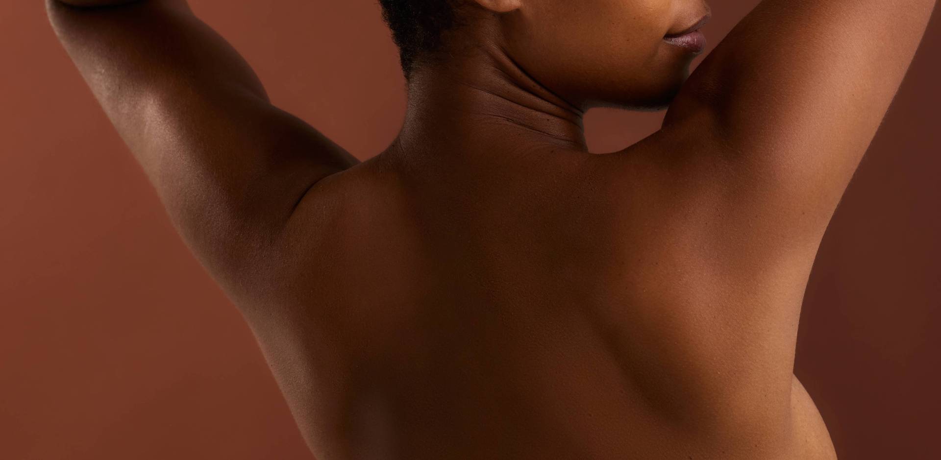 Vue de dos d'une femme noire aux cheveux courts, les bras levés derrière la tête, sur un fond marron uni.