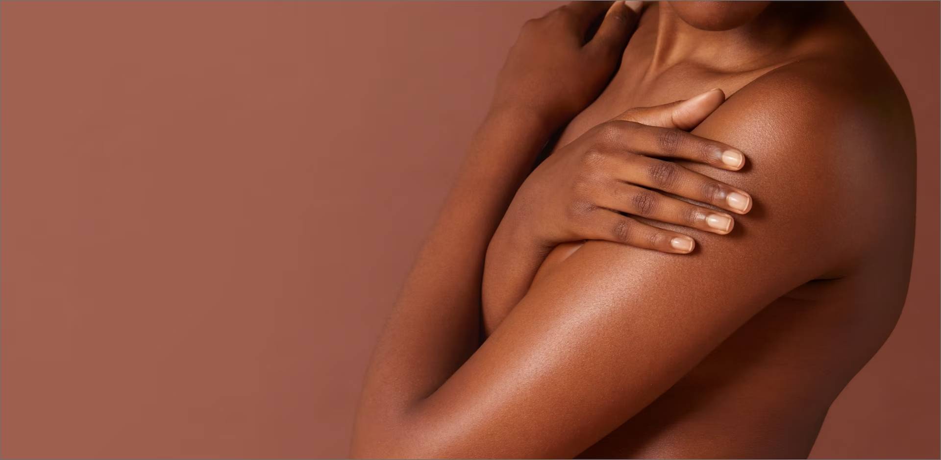 Gros plan sur le buste d'une femme à la peau foncée qui croise les bras sur sa poitrine, sur un fond marron uni.