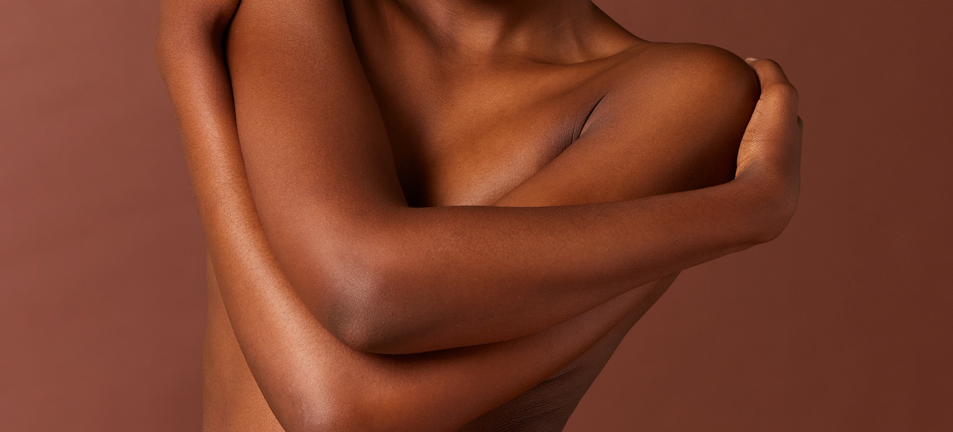 Gros plan sur le buste d'une femme à la peau foncée qui croise les bras sur sa poitrine, sur un fond marron uni.