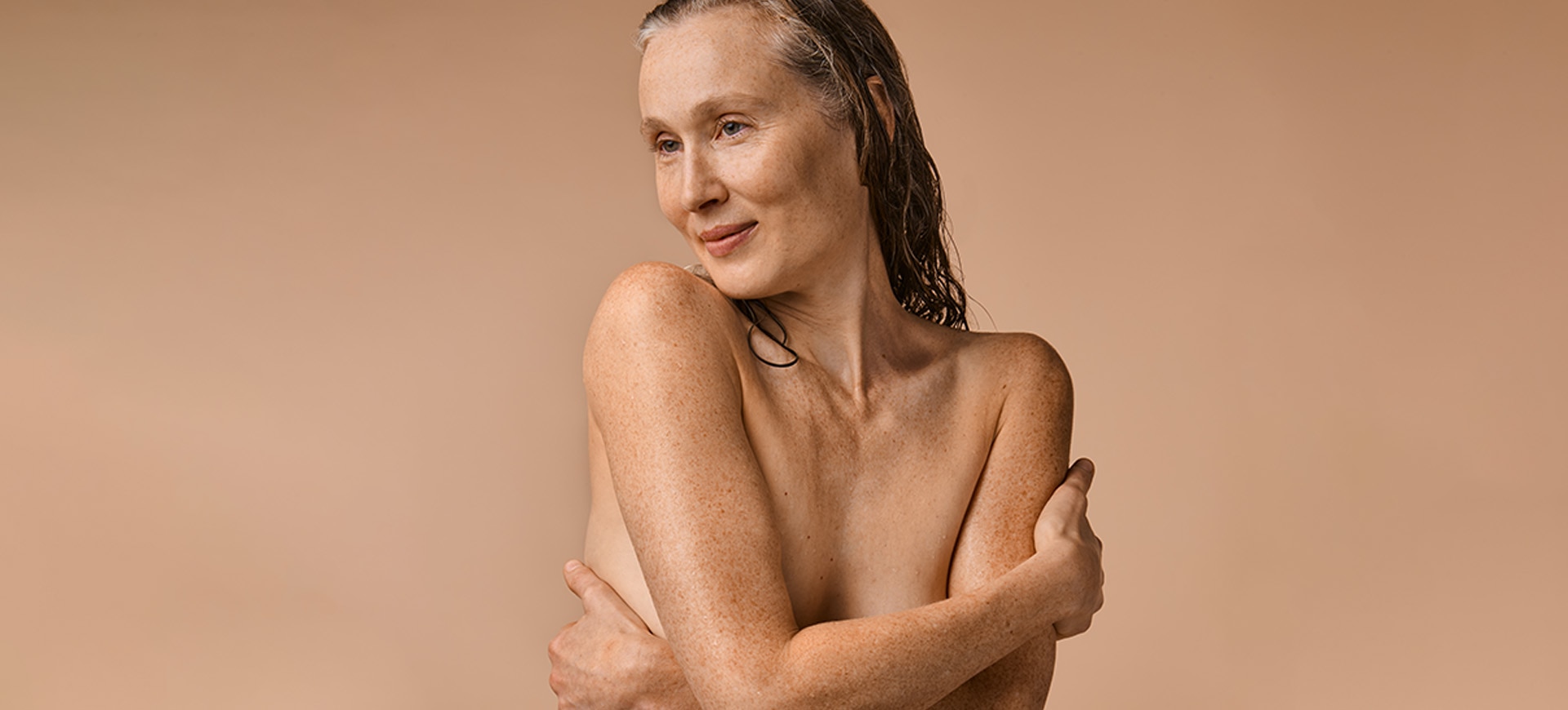 une femme âgée, fraîchement lavée, s'embrassant nue sur un fond clair de couleur chair