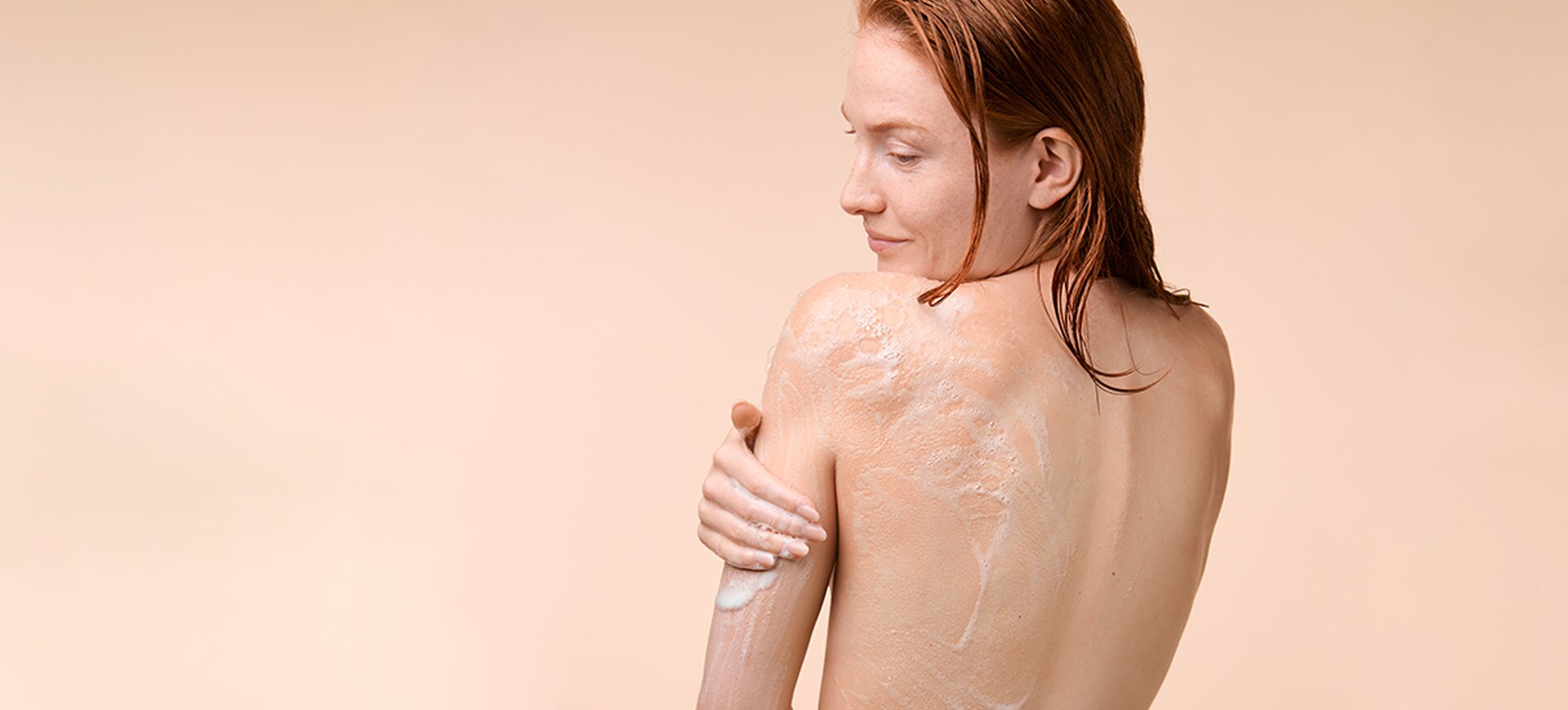 Jeune femme nue aux cheveux roux, de dos, appliquant le produit Sanex Derma Thérapie pour l'eczéma sur un fond clair de couleur chair.