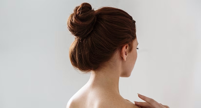 Vue de dos d'une femme rousse aux cheveux relevés en un chignon élégant. Sa main touche délicatement son épaule.