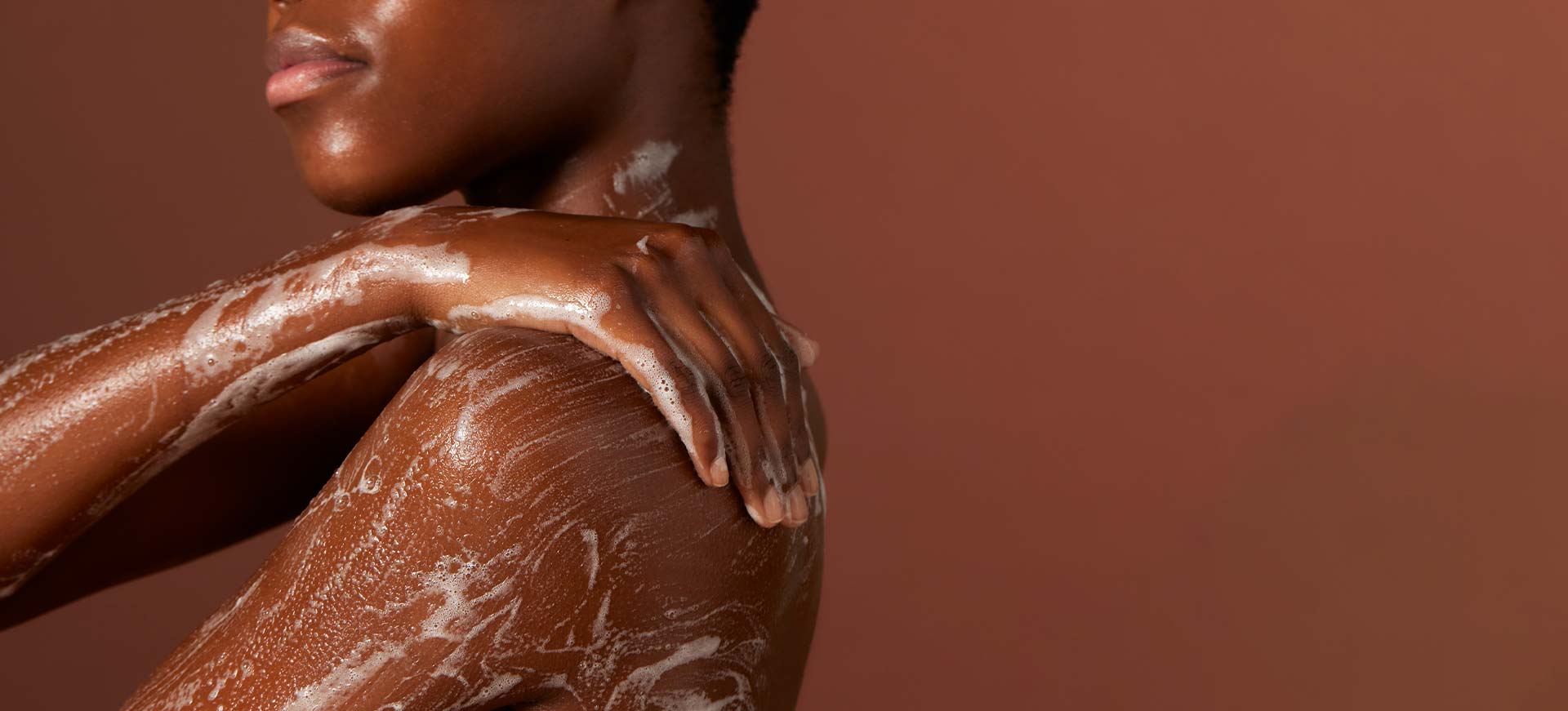 Bannière montrant une femme noire qui se savonne l'épaule et le bras avec une mousse onctueuse, sur un fond marron uni.
