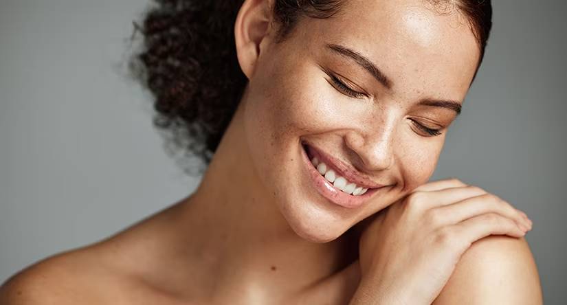 Gros plan sur le visage d'une femme souriante aux cheveux bouclés et à la peau constellée de taches de rousseur. Ses yeux sont fermés et sa main est posée sur son épaule.