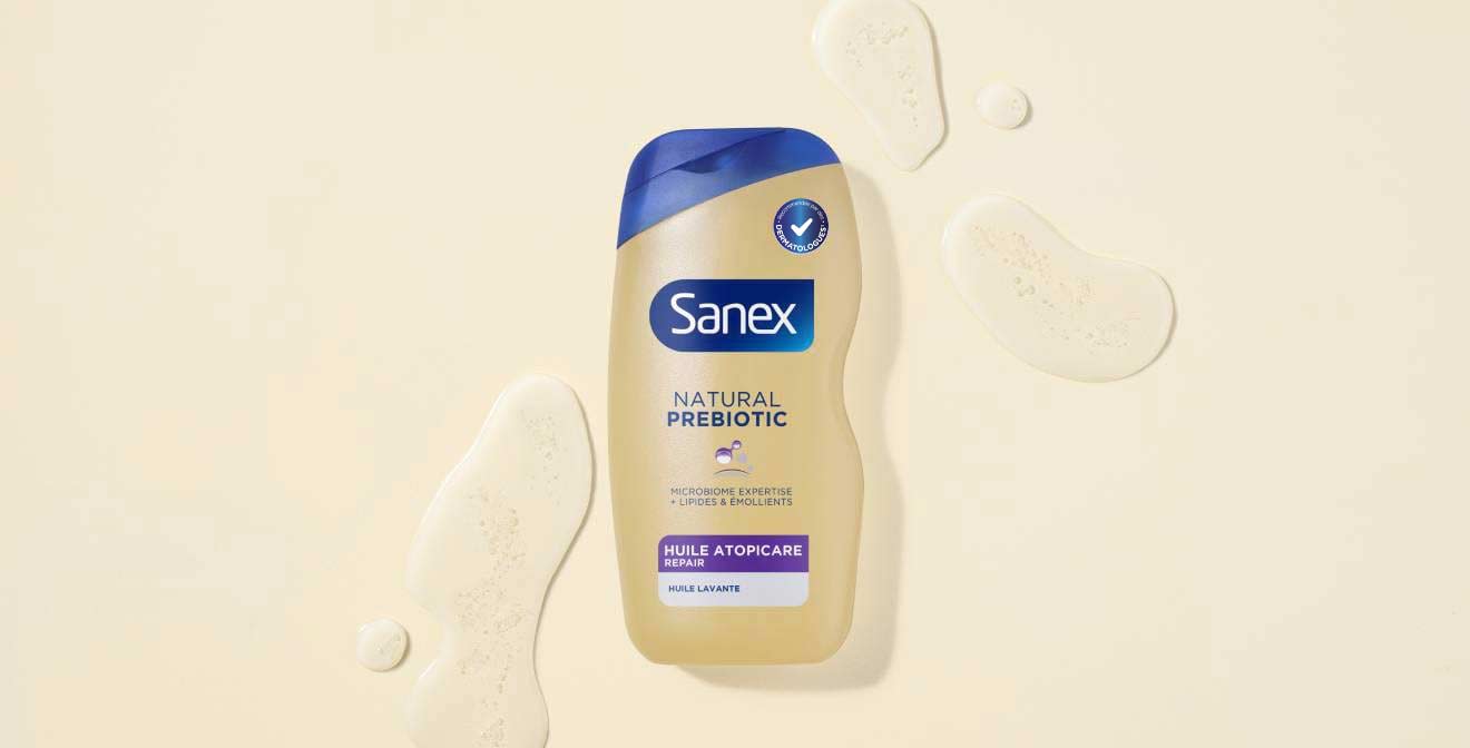 Flacon d'huile lavante Sanex Natural Prebiotic Huile Atopicare Repair, posé sur une surface beige clair. Quelques gouttes de produit s'étalent élégamment autour du flacon.