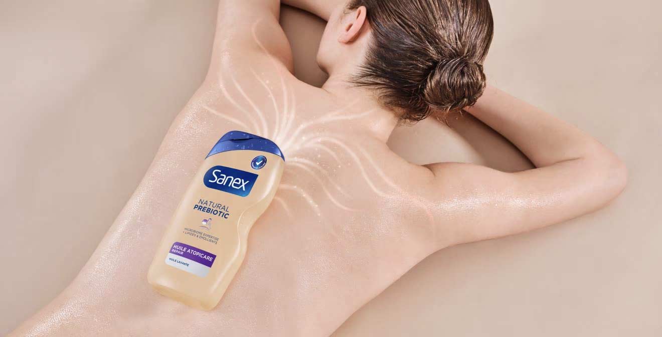 Une femme est allongée sur le ventre, le dos légèrement cambré, avec un flacon de Sanex Natural Prebiotic Huile Atopicare Repair posé sur son dos. Des lignes lumineuses symbolisent l'hydratation.