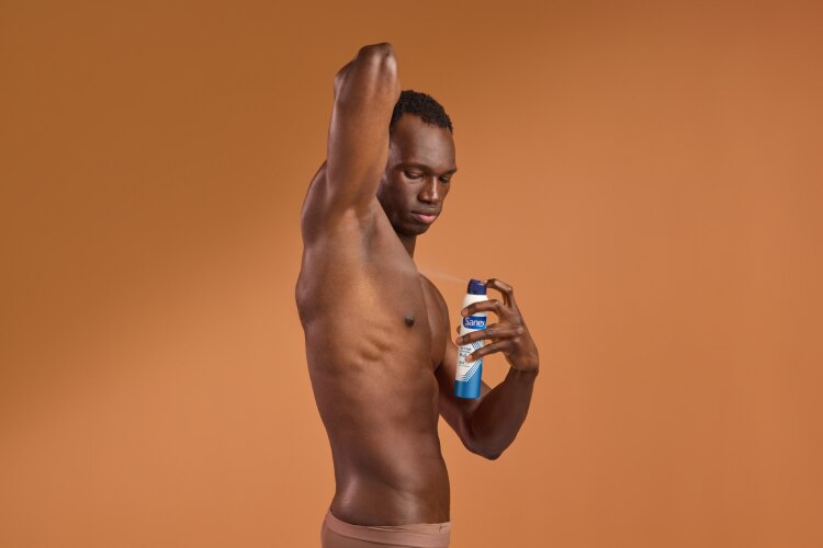Un homme noir au torse nu lève le bras pour s'appliquer un déodorant en aérosol Sanex sur son aisselle. Il est de profil sur un fond ocre uni.