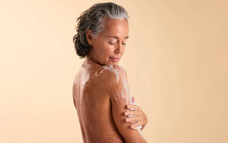 Une femme mature aux cheveux gris et ondulés se savonne l'épaule, les yeux fermés et une expression sereine.