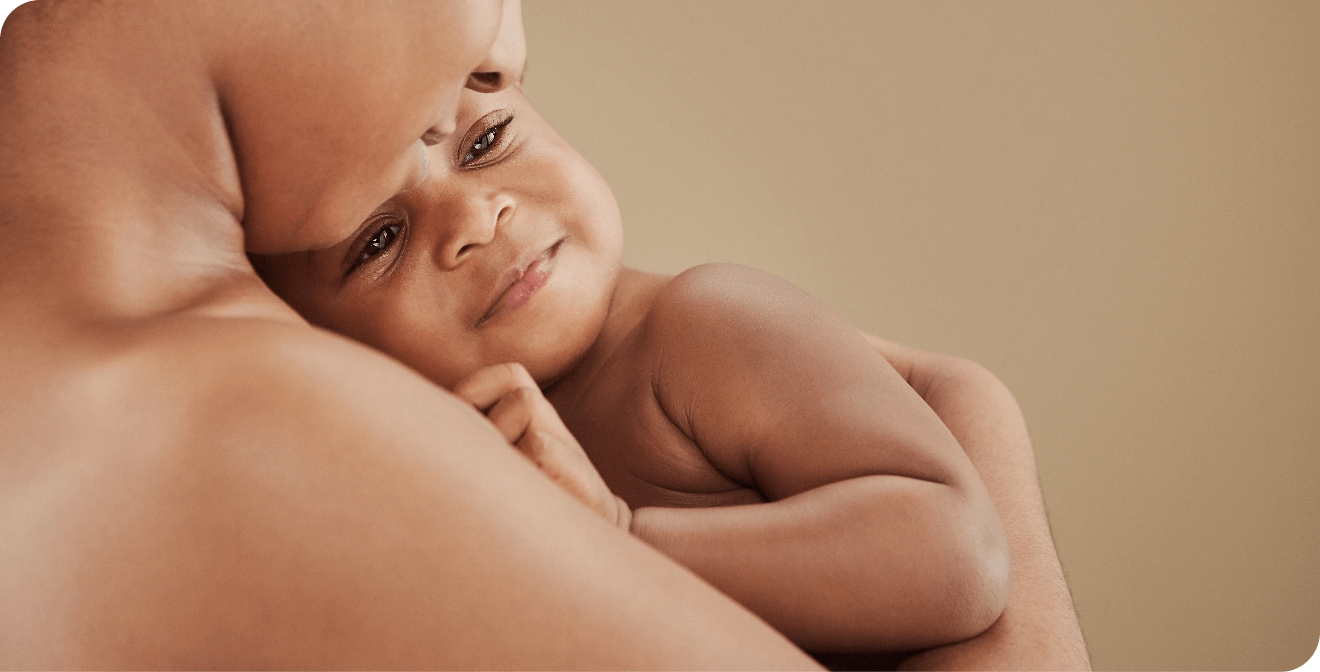 Un bébé à la peau foncée est tenu dans les bras d'un adulte, sa petite main sur sa poitrine, son visage légèrement renfrogné mais doux.