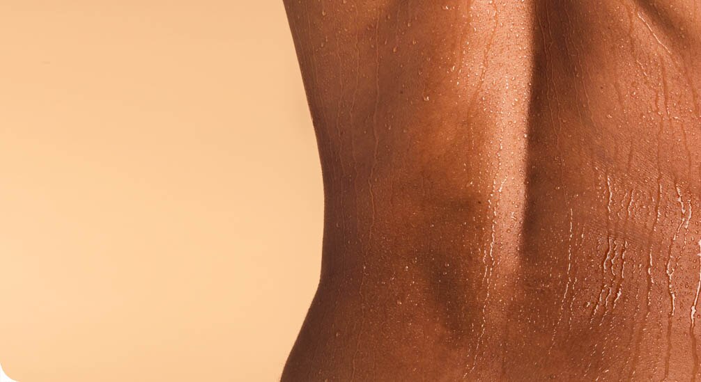 Gros plan sur le dos et le flanc d'une personne à la peau foncée et tonique, couverte de fines gouttelettes d'eau. Le fond est beige uni.