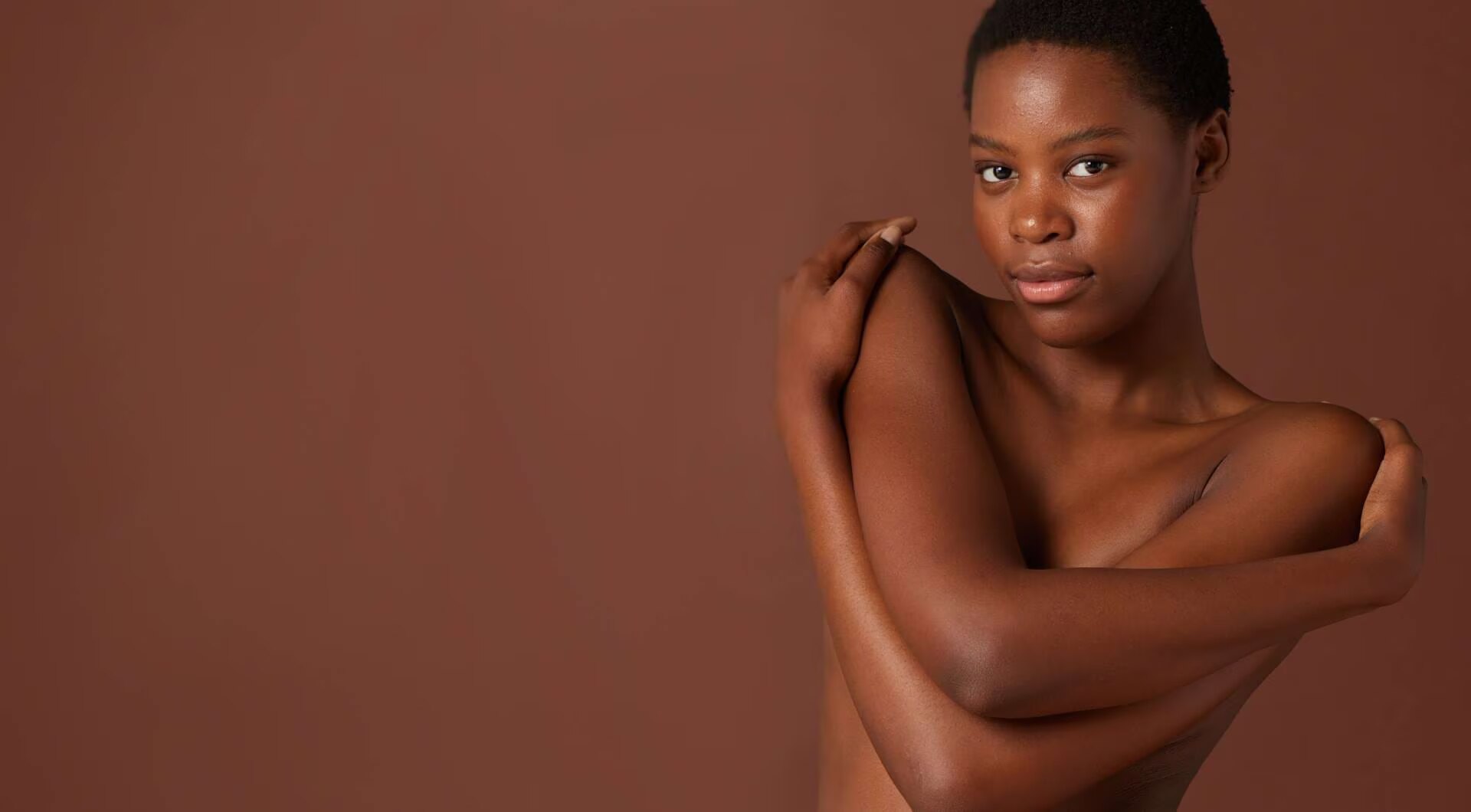 Bannière montrant une femme noire aux cheveux courts qui enlace ses épaules et regarde la caméra avec une expression douce, sur un fond marron uni.