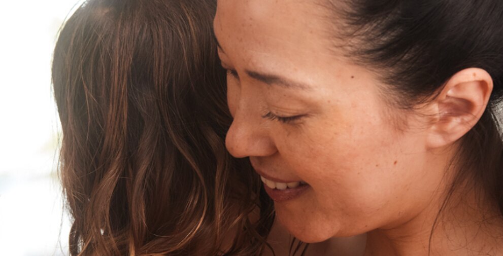 Gros plan sur le profil d'une femme souriante, les yeux fermés, qui serre tendrement dans ses bras une autre personne aux cheveux longs et ondulés.